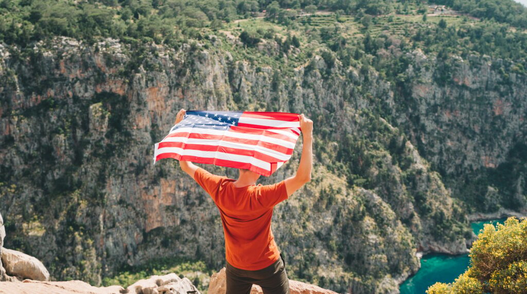 Young man standing on a rock cliff and waving the US flag while looking at sea beneath. Veteran